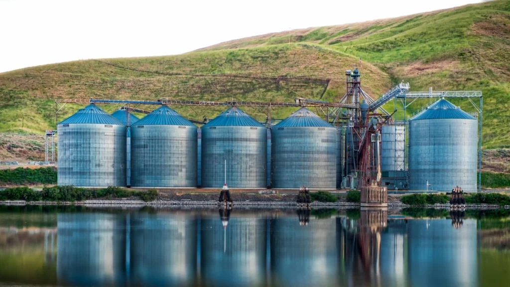 Large industrial storage silos near water with surrounding hills, representing infrastructure used in Aerobic Biological Treatment systems.